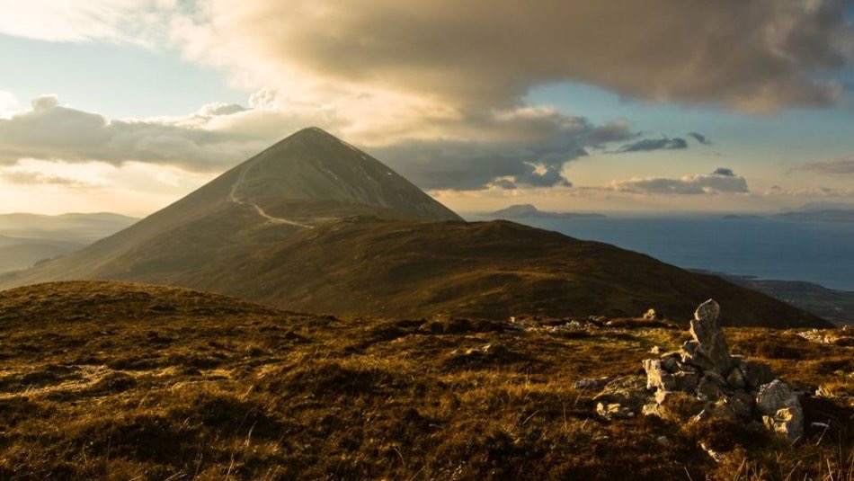 Een majestueuze berg met een opvallende piektop baadt in het warme licht van een ondergaande zon, kenmerkend voor Ierland vakanties. De voorgrond toont rotsachtig, grasachtig terrein. Wolken zweven erboven en werpen dramatische schaduwen. In de verte glinstert de zee onder een zacht verlichte hemel. | Echt Ierland Vakanties