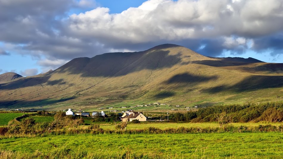 Ervaar het schilderachtige uitzicht op een weelderige groene vallei met een paar huizen, tegen glooiende heuvels onder een blauwe lucht in het serene Ierland. De heuvels en wolken werpen dramatische schaduwen, waardoor een rustige landelijke sfeer ontstaat die perfect is voor uw vakantie in Ierland. | Echt Ierland Vakanties