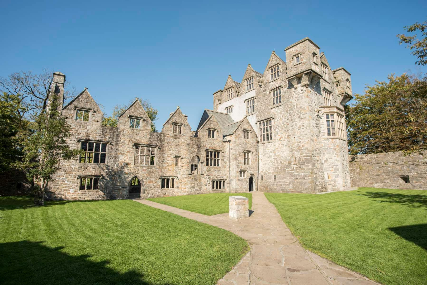 Dit historische kasteel in Ierland, gelegen op een weelderig groen gazon met kruisende stenen paden, heeft grijze stenen muren, meerdere torens en puntdaken. Zonlicht verlicht de hoge ramen en het ingewikkelde steenwerk van de gevel, waardoor het een betoverende stop is op elke rondreis door Ierland. | Echt Ierland Vakanties