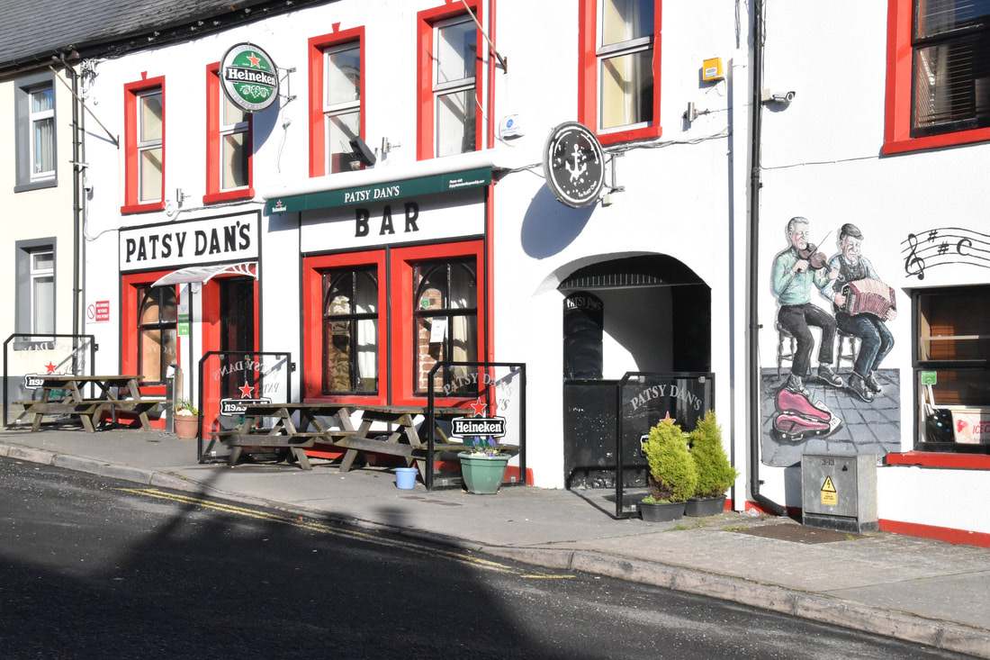 Straatbeeld van Patsy Dan's Bar, een charmante stop op uw rondreis Ierland, met rood omrande ramen en deuren. Picknicktafels staan aan de voorkant onder een groen Heineken-bord. Een muurschildering van muzikanten voegt een artistiek tintje toe aan de witte gevel, perfect voor uw vakantie in Ierland. | Echt Ierland Vakanties