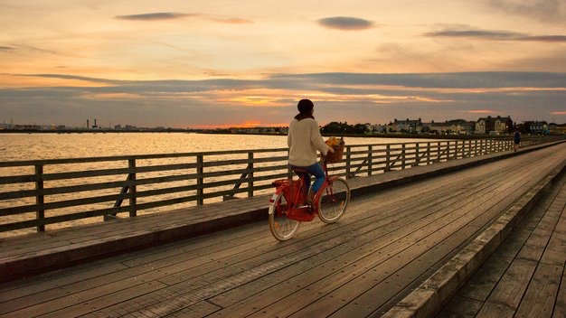 Een vrouw geniet van een rustige avond, fietsend op een houten promenade langs de waterkant bij zonsondergang tijdens haar rondreis Ierland. De lucht is versierd met warme tinten oranje en roze, weerspiegeld op het kalme water, terwijl gebouwen en een verre kustlijn bijdragen aan de serene sfeer. | Echt Ierland Vakanties