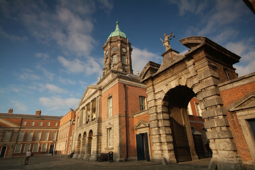 Ontdek een historisch gebouw dat doet denken aan de gebouwen die u tijdens een rondreis door Ierland ziet, met zijn centrale klokkentoren met een groene koepel die contrasteert met de blauwe lucht. Gemaakt van rode baksteen en steen, het heeft boogramen en zuilen. Een stenen boog met een standbeeld erop leidt naar een charmante binnenplaats aan de linkerkant. | Echt Ierland Vakanties