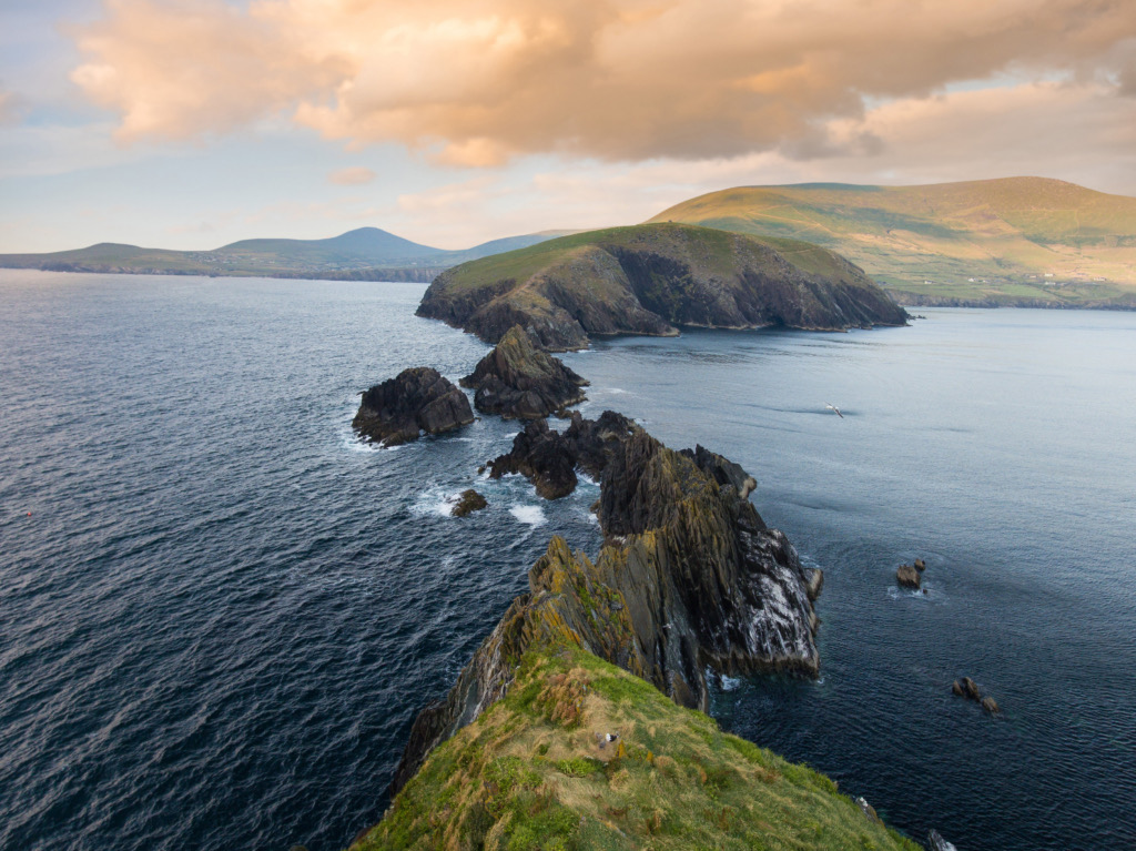 Ervaar een schilderachtig uitzicht op de ruige kustlijn van Ierland, waar grillige kliffen zich uitstrekken tot in de zee onder een hemel met verspreide wolken en een warme gloed van de zonsondergang. Groene heuvels rijzen op de achtergrond en golven klotsen zachtjes tegen de kliffen, wat een sereen en schilderachtig landschap biedt tijdens uw vakantie in Ierland. | Echt Ierland Vakanties