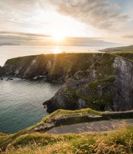 Ervaar een schilderachtig uitzicht op de kust bij zonsondergang tijdens uw vakantie in Ierland, waar ruige kliffen bedekt met groene vegetatie uitkijken over een kalme oceaan. Een kronkelend pad volgt de rand van de klif tot aan de horizon, met verre eilanden zichtbaar onder de wolkenlucht tijdens uw Ierland-tour. | Echt Ierland Vakanties