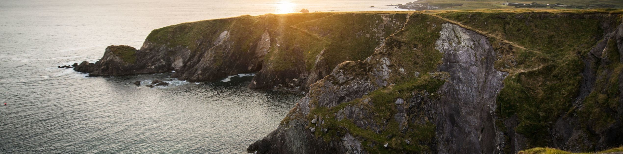 Ervaar een schilderachtig uitzicht op de kust bij zonsondergang tijdens uw vakantie in Ierland, waar ruige kliffen bedekt met groene vegetatie uitkijken over een kalme oceaan. Een kronkelend pad volgt de rand van de klif tot aan de horizon, met verre eilanden zichtbaar onder de wolkenlucht tijdens uw Ierland-tour. | Echt Ierland Vakanties