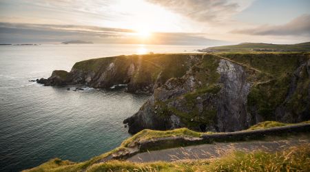 Ervaar een schilderachtig uitzicht op de kust bij zonsondergang tijdens uw vakantie in Ierland, waar ruige kliffen bedekt met groene vegetatie uitkijken over een kalme oceaan. Een kronkelend pad volgt de rand van de klif tot aan de horizon, met verre eilanden zichtbaar onder de wolkenlucht tijdens uw Ierland-tour. | Echt Ierland Vakanties