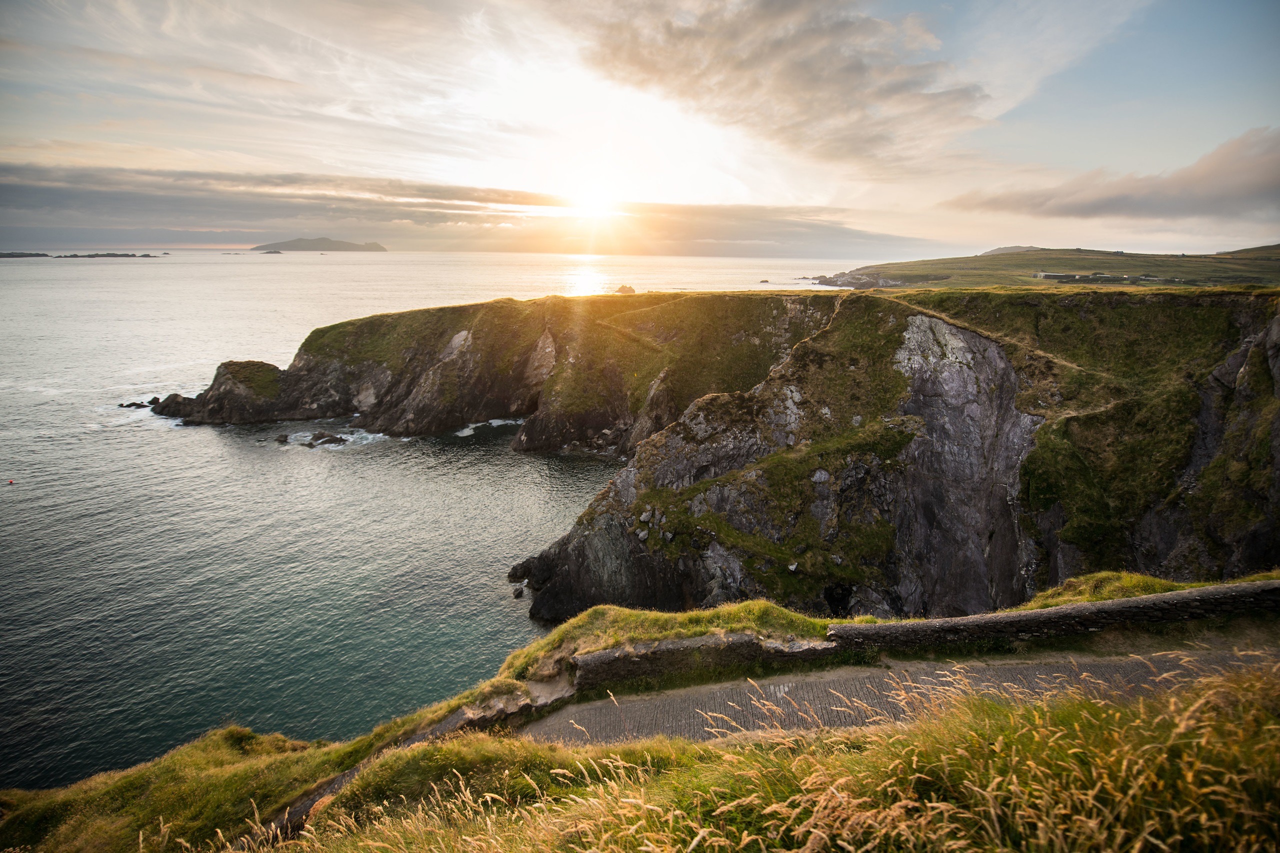 Ervaar een schilderachtig uitzicht op de kust bij zonsondergang tijdens uw vakantie in Ierland, waar ruige kliffen bedekt met groene vegetatie uitkijken over een kalme oceaan. Een kronkelend pad volgt de rand van de klif tot aan de horizon, met verre eilanden zichtbaar onder de wolkenlucht tijdens uw Ierland-tour. | Echt Ierland Vakanties