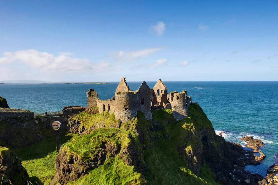 Gelegen op weelderige groene kliffen, kijken de oude stenen ruïnes van Dunluce Castle uit over de diepblauwe zee, een must-see op elke rondreis Ierland. Golven beuken tegen de rotsachtige kustlijn beneden, terwijl een heldere lucht met wolken zich uitstrekt boven, waardoor het een onvergetelijke stop is op uw vakantie Ierland. | Echt Ierland Vakanties