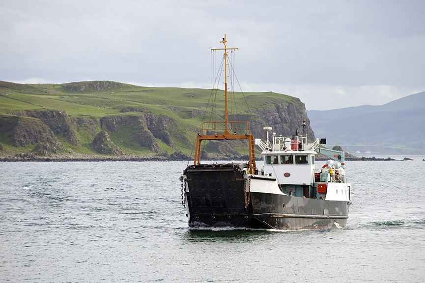 Een kleine veerboot met een zwarte romp vaart over kalme wateren langs een ruige kustlijn, perfect voor degenen die genieten van een vakantie in Ierland. Het land heeft groene, met gras begroeide kliffen en glooiende heuvels onder een bewolkte lucht. Mensen zijn te zien op het dek van de veerboot, die naar de kijker toe bewegen. | Echt Ierland Vakanties