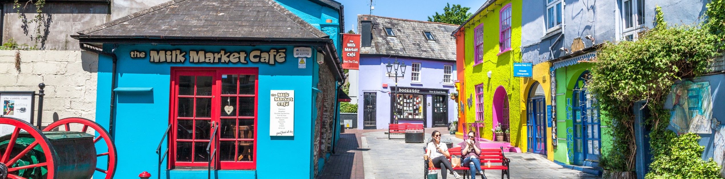 Een levendig straatbeeld in een schilderachtig Iers dorp met kleurrijke gebouwen, waaronder het felblauwe "The Milk Market Café". Bezoekers van rondreis Ierland loungen op bankjes langs de geplaveide straat. Een grote kar en paaltjes maken dit charmante tafereel, geschilderd in blauw, geel en paars, compleet. | Echt Ierland Vakanties
