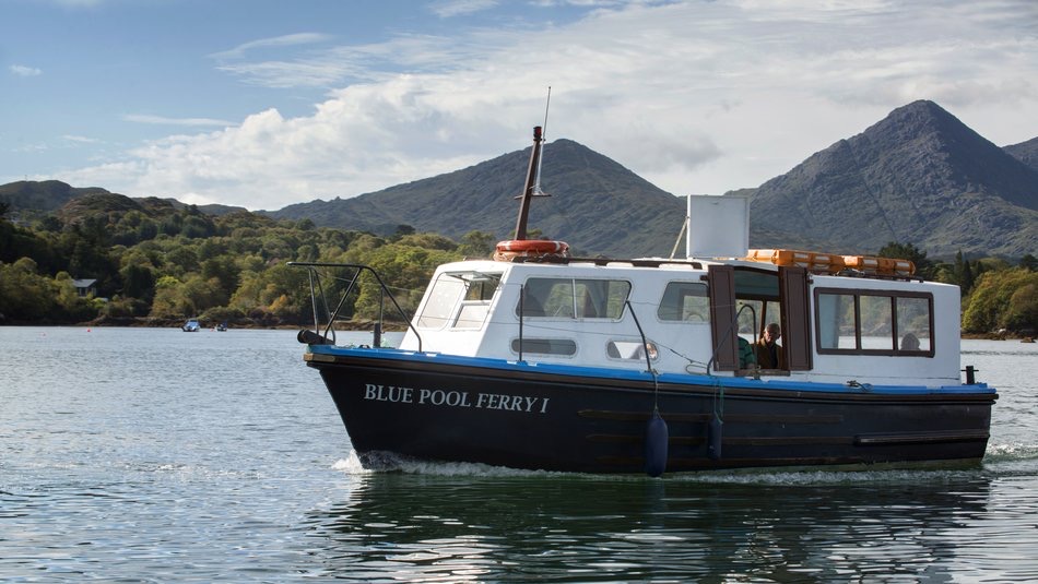 De kleine veerboot, "Blue Pool Ferry I," glijdt over een kalm water. De witte cabine met blauwe en zwarte afwerking complementeert de weelderige groene heuvels en grillige bergen onder een gedeeltelijk bewolkte lucht—perfect voor degenen die een onvergetelijke rondreis Ierland maken. | Echt Ierland Vakanties