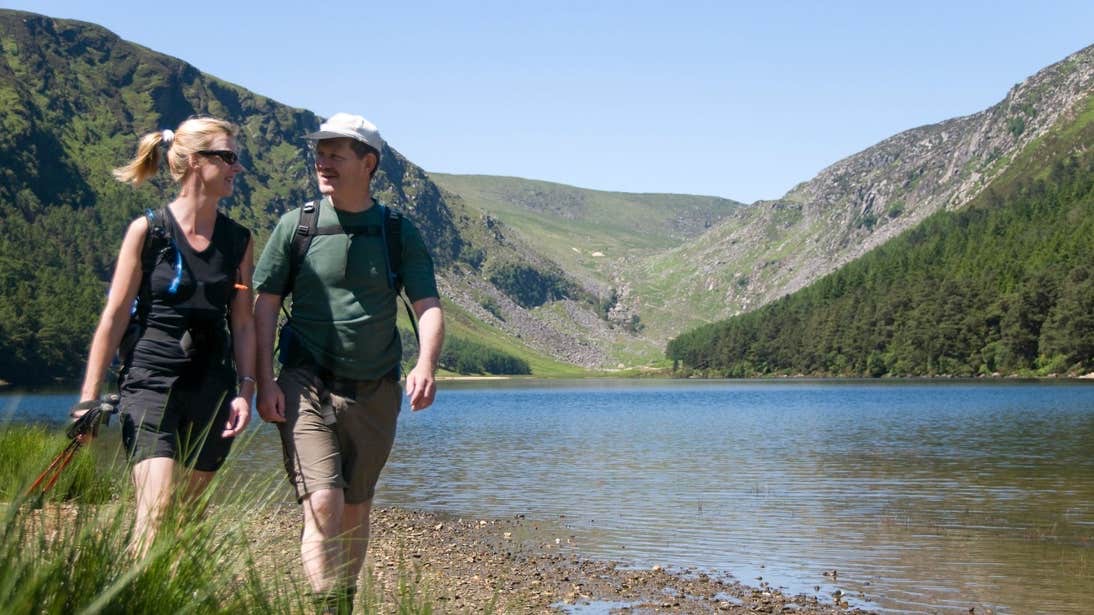 Een stel geniet van een rustige wandeling langs de oever van een rustig meer, omlijst door weelderige groene heuvels en bergen in het schilderachtige Ierland. De vrouw, in een zwarte outfit en zonnebril, en de man, in een groen shirt en pet, koesteren zich onder de helderblauwe lucht op hun onvergetelijke Ierland vakantie. | Echt Ierland Vakanties