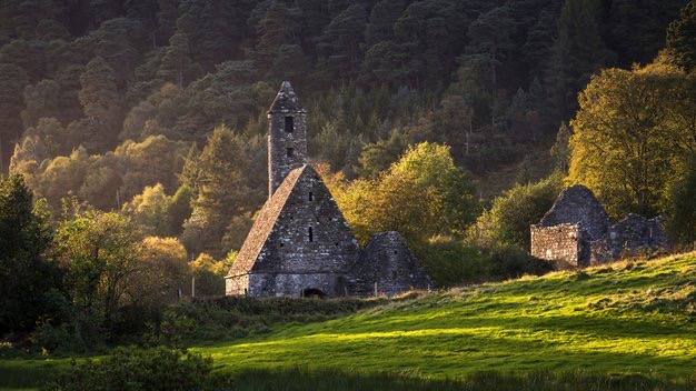 Een serene scène van een oude stenen kerk met een hoge, puntige klokkentoren staat tegen een dichte bosachtergrond. De zon werpt warm licht op de kerk en het weelderige groen, ideaal voor degenen die op zoek zijn naar een vakantie in Ierland. Een klein stenen bouwwerk is in de buurt zichtbaar. | Echt Ierland Vakanties