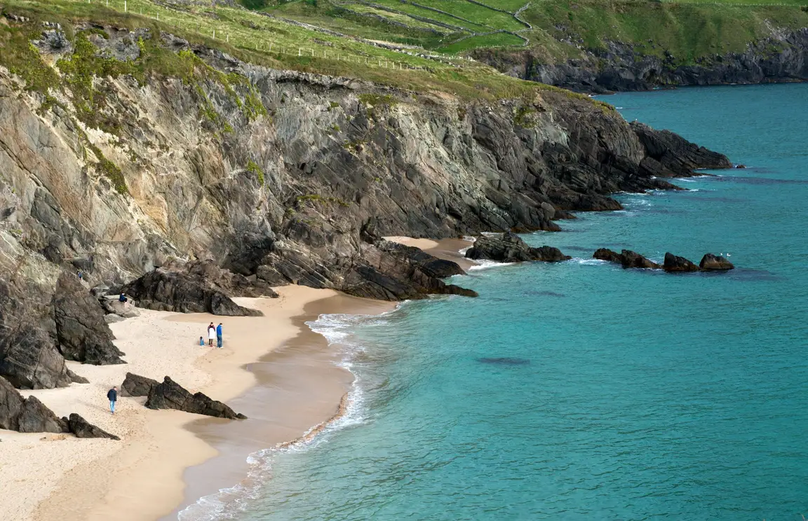 Een schilderachtige kustfoto legt de essentie van een vakantie in Ierland vast, met een rotsachtige klif versierd met stukken groen die een zandstrand ontmoeten. De turquoise oceaan klotst zachtjes tegen de kust terwijl een groep mensen bij de kliffen staat en geniet van het serene landschap dat kenmerkend is voor vakanties in Ierland. | Echt Ierland Vakanties