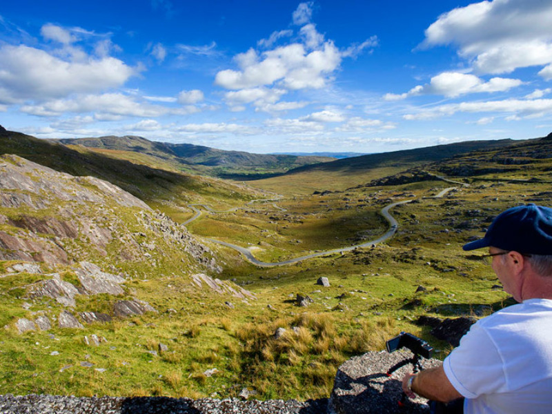 Een persoon in een wit shirt en blauwe pet filmt een schilderachtig landschap, en legt de essentie van een rondreis Ierland vast. Het uitzicht toont glooiende groene heuvels, een kronkelende weg en verspreide rotsen onder een helderblauwe lucht met donzige wolken. Perfect voor degenen die dromen van bijzondere Ierland vakanties. | Echt Ierland Vakanties