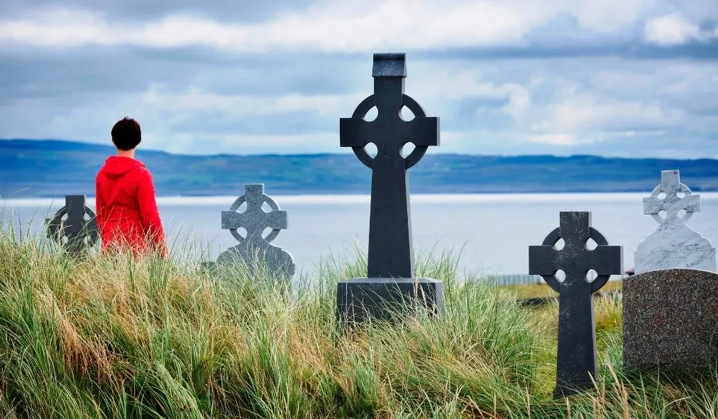 Een persoon in een rood jasje staat tussen de met gras bedekte duinen, met het gezicht naar meerdere Keltische kruisgrafstenen. Het tafereel, dat doet denken aan een serene vakantie in Ierland, kijkt uit over een kalm waterlichaam onder een bewolkte lucht, wat een contemplatieve sfeer creëert. | Echt Ierland Vakanties