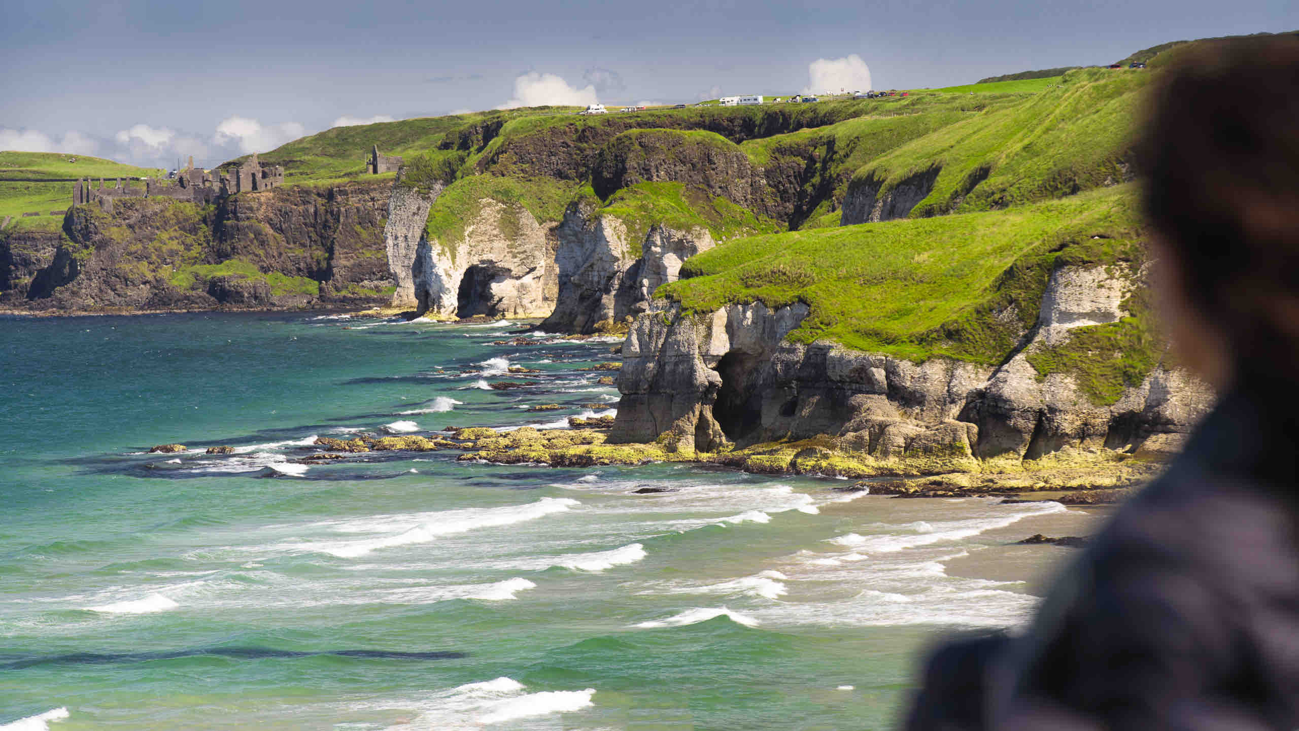 Een persoon kijkt uit over een schilderachtig kustlandschap tijdens hun vakantie in Ierland, waar ruige kliffen en rotsachtige uitsteeksels smaragdgroene kliffen ontmoeten die dramatisch oprijzen uit turquoise golven. De heldere lucht houdt een paar wolken vast en oude ruïnes zijn zichtbaar bovenop de kliffen in deze onvergetelijke rondreis Ierland. | Echt Ierland Vakanties