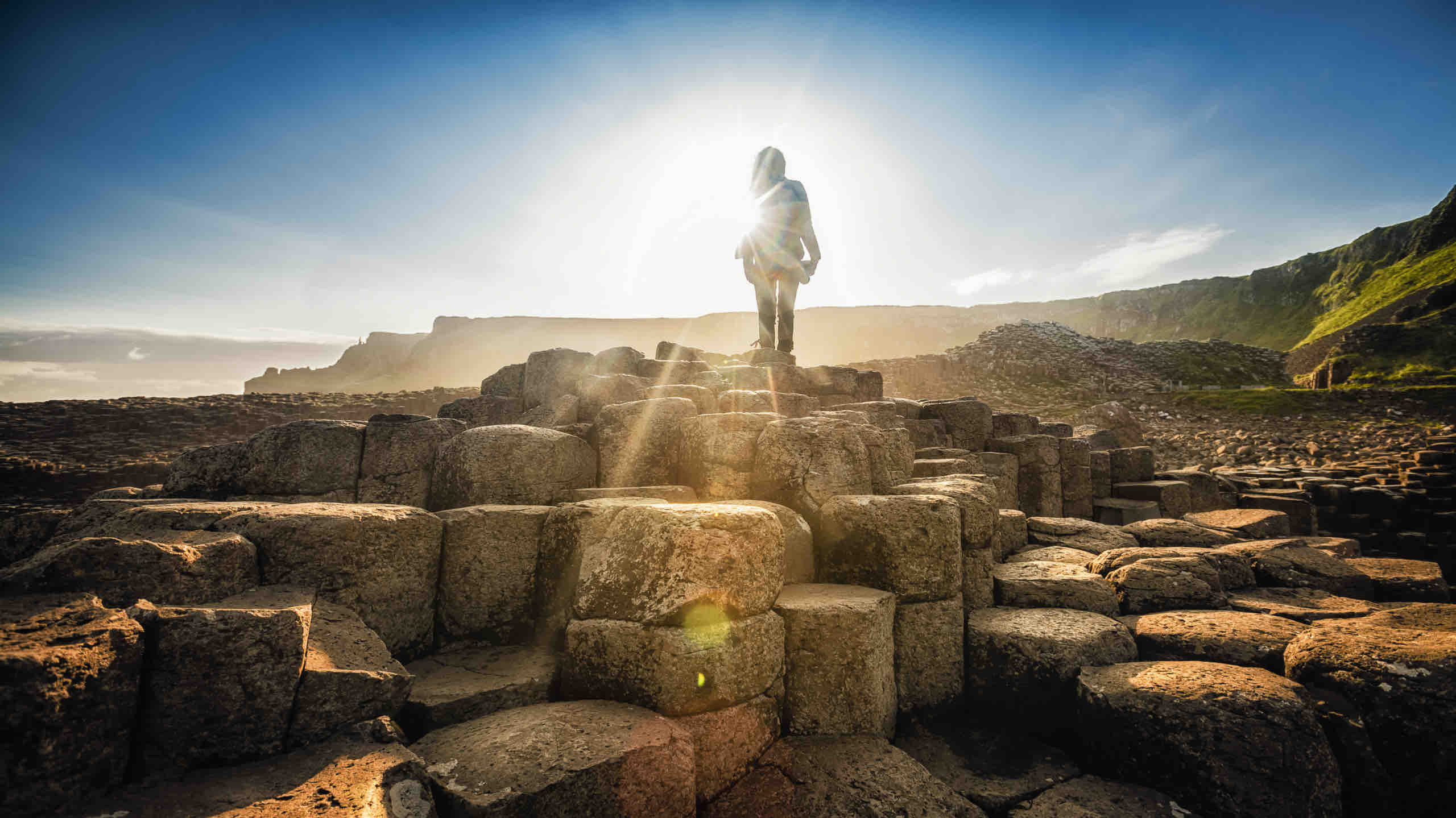 Een persoon staat bovenop de Giant's Causeway, gesilhouetteerd door een felle zon die een halo-effect creëert. De basaltkolommen vormen een uniek, geometrisch landschap, perfect voor rondreis Ierland. Ruige kliffen en een helderblauwe lucht accentueren de natuurlijke schoonheid van deze prachtige vakantiebestemming in Ierland. | Echt Ierland Vakanties