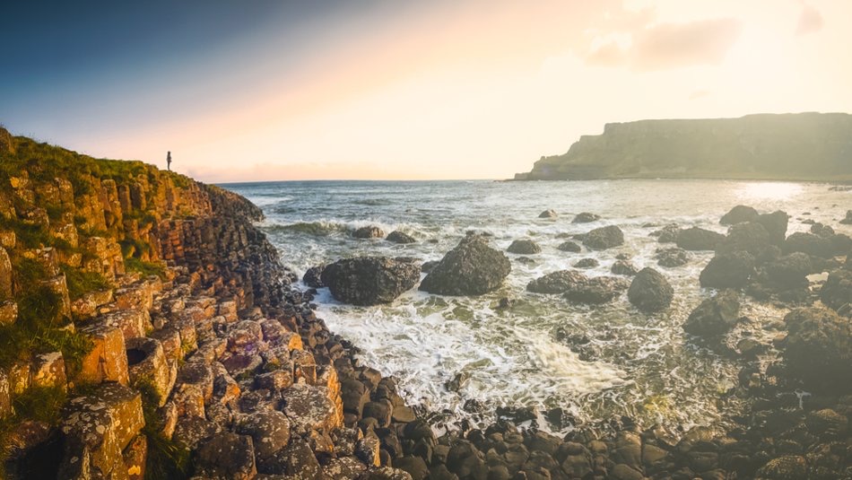 Een kustlandschap bij zonsopgang, perfect voor een rondreis Ierland, toont hexagonale basaltkolommen op de voorgrond. De rotsachtige kust ontmoet oceaan golven die tegen rotsblokken beuken. Gouden wolken vullen de lucht en een verre klif doemt op terwijl een reiziger links staat en het adembenemende uitzicht bewondert. | Echt Ierland Vakanties