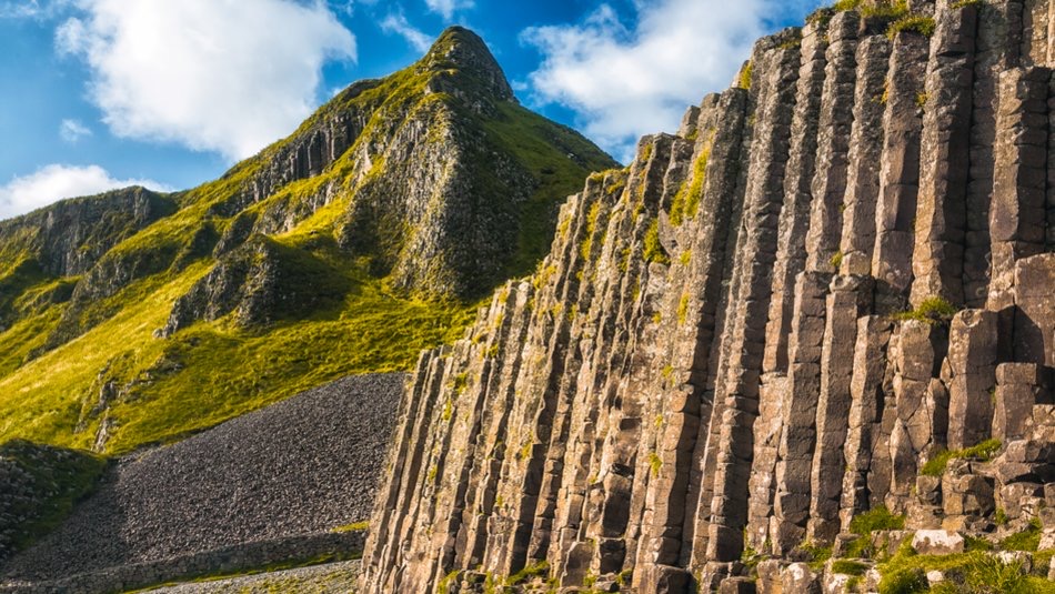 Zonovergoten basaltkolommen vormen een opvallende geologische formatie, hoog oprijzend tegen een ruige, met gras begroeide berghelling - een perfecte momentopname van uw rondreis Ierland. De lucht is helder met verspreide wolken, die zachte schaduwen werpen op het landschap en bijdragen aan de natuurlijke schoonheid van dit unieke tafereel. | Echt Ierland Vakanties