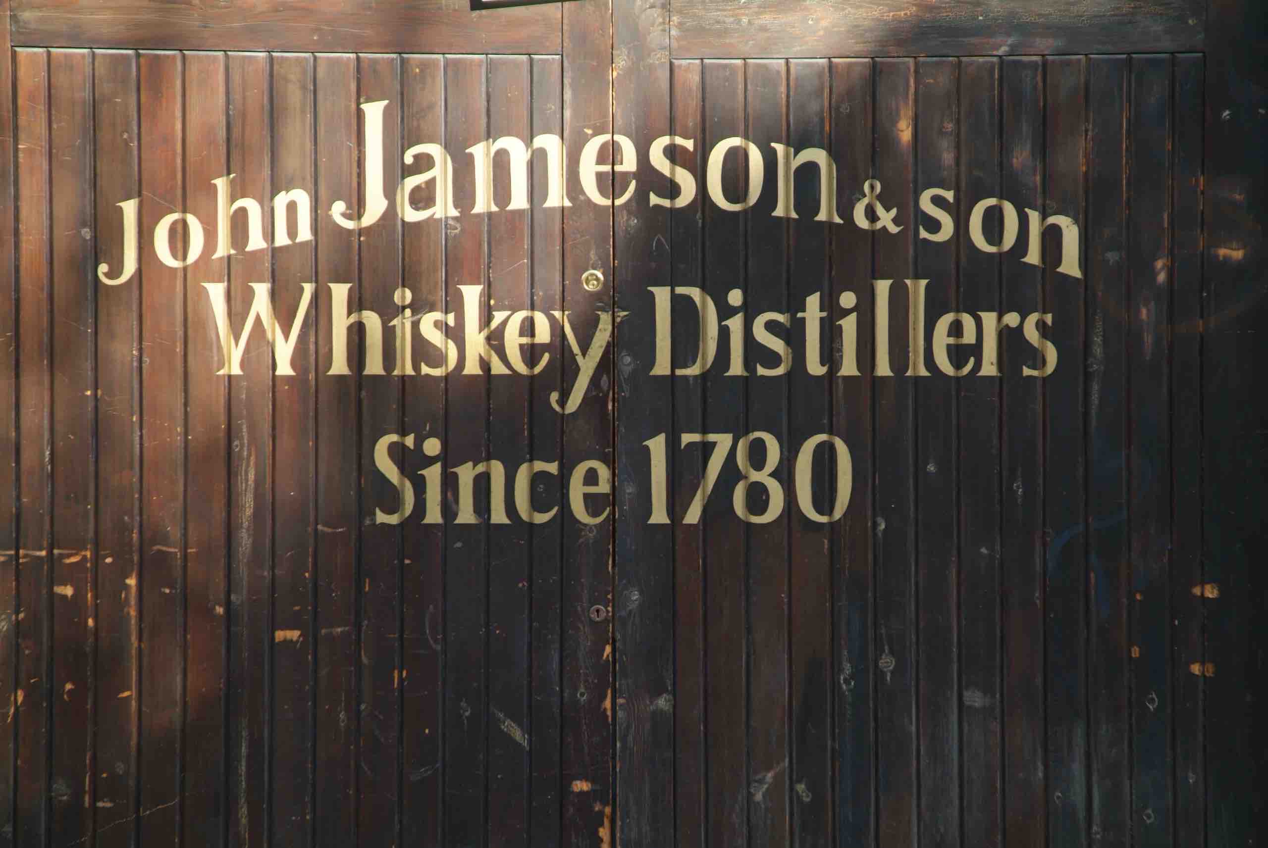 Een verweerde houten deur in Ierland, met afbladderende verf en gouden letters met de tekst "John Jameson & Son Whiskey Distillers Since 1780", roept de charme op van een Ierse vakantie. Het licht gebogen, versleten oppervlak met zijn krassen creëert een rustieke allure die perfect is voor een rondreis Ierland. | Echt Ierland Vakanties