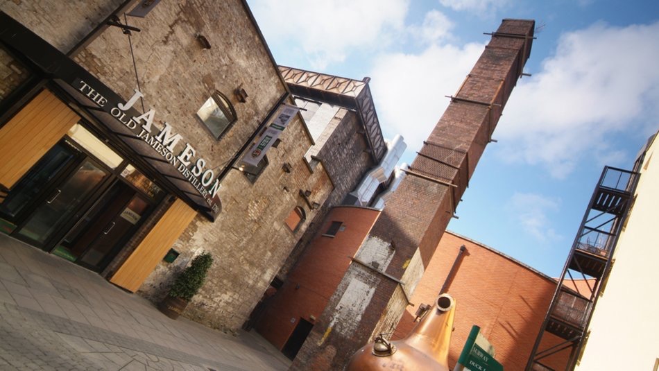 De afbeelding legt de buitenkant vast van de Old Jameson Distillery, een must-see op elke rondreis door Ierland, met een rustiek bakstenen gebouw met een hoge schoorsteen en industriële structuren. Een koperen pot staat prominent op de voorgrond onder een blauwe lucht bezaaid met wolken. | Echt Ierland Vakanties
