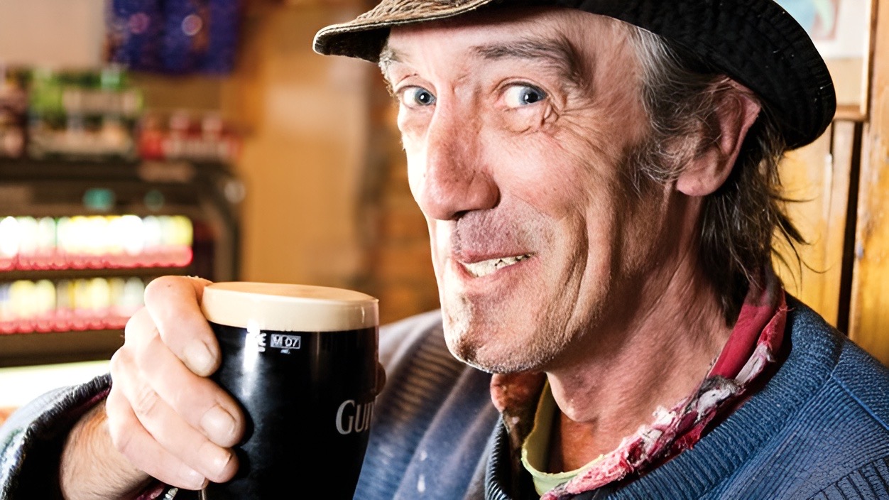 Een man met een hoed en sjaal lacht hartelijk terwijl hij een pint Guinness vasthoudt in een bar, wat de geest van rondreis Ierland belichaamt. Vage planken vol met flessen creëren een gezellige sfeer, die de essentie van Ierland vakanties perfect vastlegt in zijn relaxte, casual kleding. | Echt Ierland Vakanties