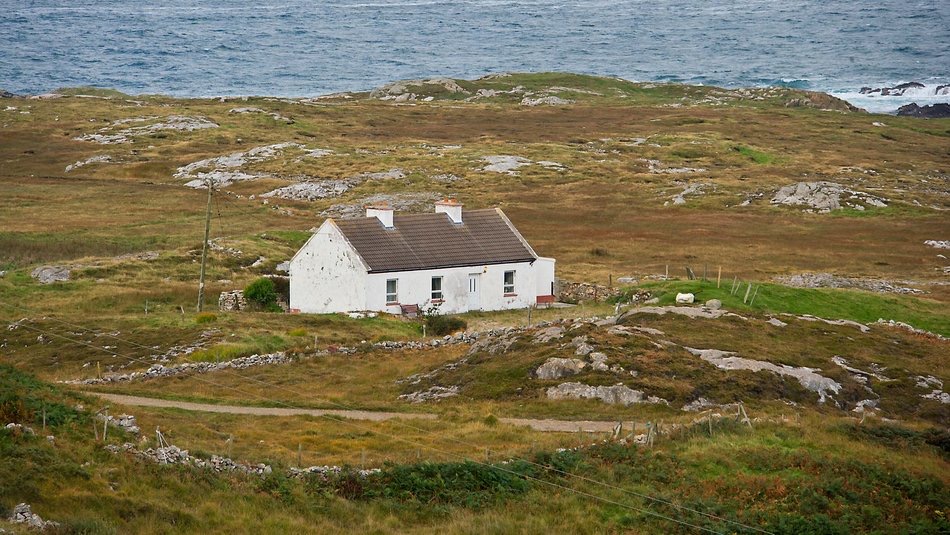 Een eenzaam wit huis met een donker dak staat in een uitgestrekt, rotsachtig landschap met schaars gras en een kronkelend zandpad. De oceaan is in de verte zichtbaar onder een bewolkte lucht. Stenen ruïnes en stukken groen omringen het gebied, wat de afgelegen rust creëert die perfect is voor een rondreis door Ierland. | Echt Ierland Vakanties