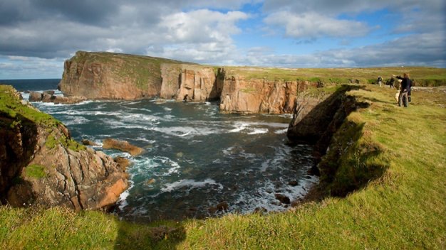 Een schilderachtig kustgezicht in Ierland toont ruige kliffen en een rotsachtige kustlijn. Het groene graslandschap contrasteert met de roodbruine kliffen terwijl de oceaan golven beuken onder een gedeeltelijk bewolkte lucht. Een paar mensen staan op de rand en genieten van het uitzicht - een perfecte plek voor een Ierland vakantie-ervaring. | Echt Ierland Vakanties