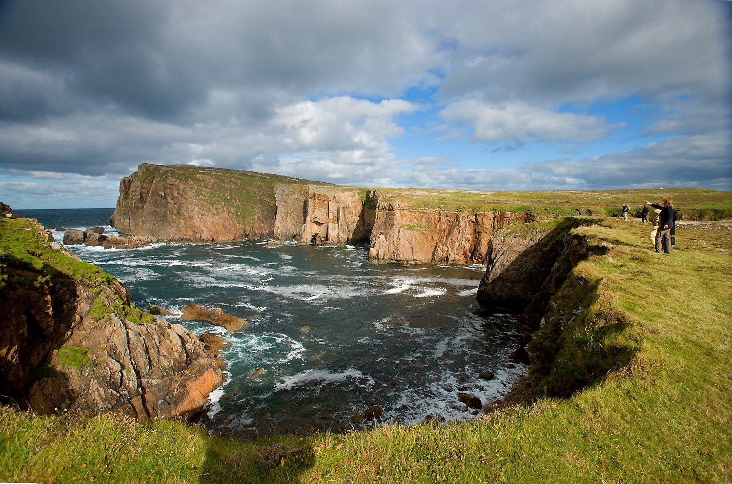 Ervaar het ruige kustlandschap van Ierland, waar steile kliffen een rotsachtige kustlijn ontmoeten onder een gedeeltelijk bewolkte lucht. Oceaan golven beuken tegen de kliffen terwijl een paar mensen langs de rand lopen, allemaal verlicht door zacht, natuurlijk licht - perfect voor een onvergetelijke vakantie in Ierland. | Echt Ierland Vakanties