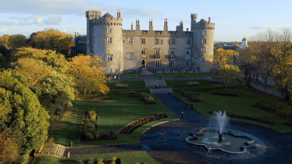 Luchtfoto van Kilkenny Castle omlijst door herfstbomen tijdens een onvergetelijke vakantie Ierland. Het stenen kasteel heeft ronde torens en kantelen met kantelen. Beneden siert een fontein een ronde vijver te midden van verzorgde gazons. Bezoekers op een rondreis Ierland genieten van de serene tuinpaden. | Echt Ierland Vakanties
