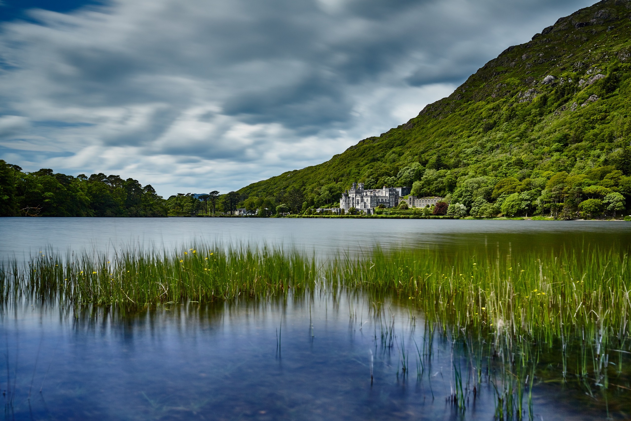 Een schilderachtig uitzicht op het meer in het vakantie-perfecte Ierland toont een historisch stenen gebouw genesteld tussen weelderig groen en heuvels. De voorgrond toont hoog gras dat groeit in het kalme water, terwijl een bewolkte lucht zich uitstrekt boven het water, wat een serene sfeer creëert die ideaal is voor uw vakantie in Ierland. | Echt Ierland Vakanties