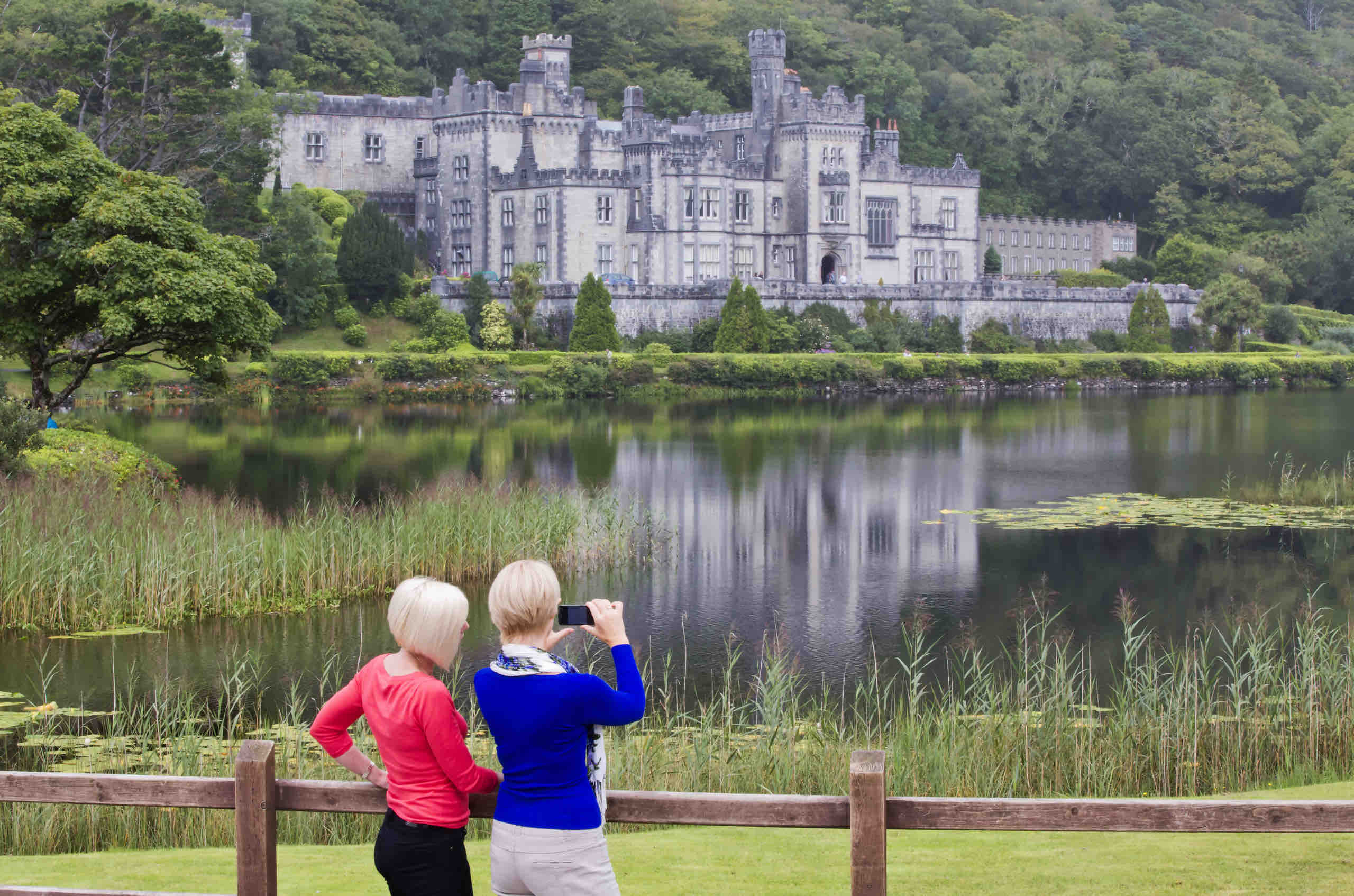 Twee vrouwen bewonderen een groot kasteel aan de overkant van een meer tijdens hun vakantie in Ierland, omringd door weelderig groen. De weerspiegeling van het kasteel schittert op het water. Een vrouw in het rood wijst terwijl een andere in het blauw toekijkt. Hoog gras grenst aan het kalme meer en dichte bomen rijzen op achter het kasteel. Een houten hek loopt op de voorgrond. | Echt Ierland Vakanties