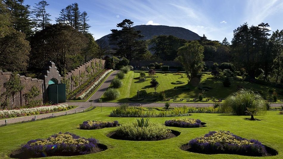 Een weelderige tuin met verzorgde gazons, symmetrische bloemperken en kronkelende paden die doen denken aan een betoverende vakantie in Ierland. Een hoge bakstenen muur omzoomt de linkerkant met kleurrijke bloemen. Op de achtergrond maken dichte bomen en een hoge berg onder een blauwe lucht met dunne wolken het rustige landschap compleet. | Echt Ierland Vakanties