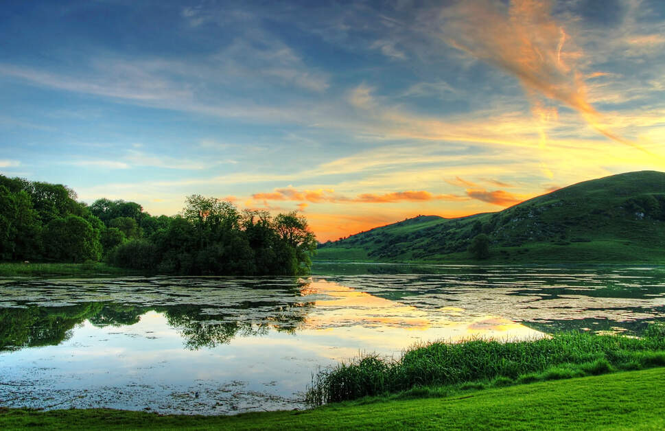 Een sereen meer weerspiegelt de levendige kleuren van een zonsondergang, met tinten oranje en blauw in de lucht. Het water is omgeven door weelderig groen gras en struiken. Een heuvel rijst op aan de rechterkant en dunne wolken strekken zich uit over de lucht, wat bijdraagt aan dit rustige tafereel dat doet denken aan een idyllische vakantie in Ierland. | Echt Ierland Vakanties