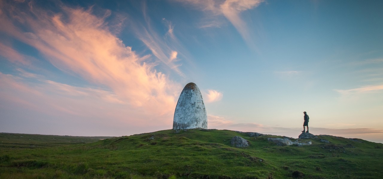 Een eenzame persoon staat op een grasheuvel met een rotsachtige richel en kijkt naar een groot, verweerd, wit monument in de vorm van een kegel. De lucht is versierd met roze en oranje wolkjes, wat een vredig en uitgestrekt sfeervol decor creëert - een idyllisch tafereel dat typerend is voor een betoverende vakantie in Ierland. | Echt Ierland Vakanties