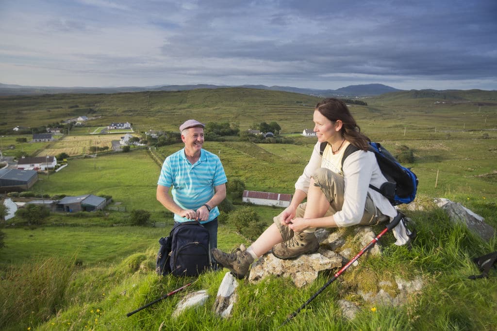 Een man en een vrouw rusten uit op een grasheuvel met wandelspullen tijdens hun rondreis Ierland. De vrouw zit op rotsen, terwijl de man op het gras zit naast een rugzak. Groene velden, verspreide huizen en een verre berg onder een bewolkte lucht verbeteren hun uitzicht. | Echt Ierland Vakanties