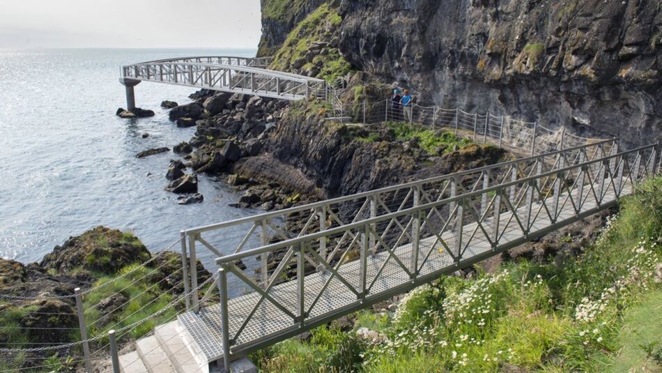 Een kustpad met twee metalen loopbruggen slingert rond rotsachtige kliffen langs de zee, perfect voor degenen die een rondreis Ierland maken. Groen gras en wilde bloemen omzomen het pad. De oceaan is kalm, met een paar mensen op de bovenste brug die genieten van het schilderachtige uitzicht op een zonnige dag tijdens hun vakantie Ierland. | Echt Ierland Vakanties