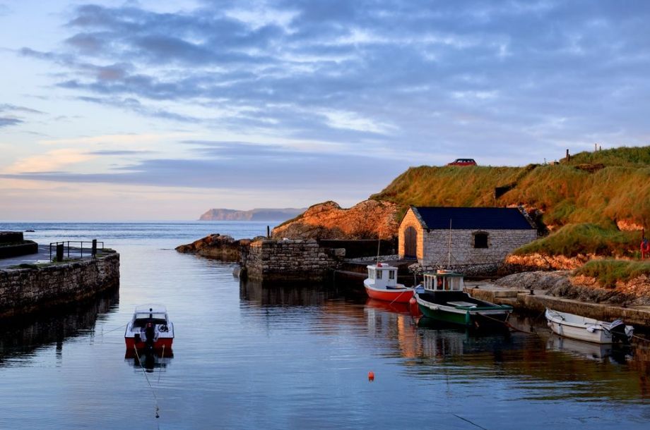Een rustig havengezicht bij zonsondergang met kleine boten afgemeerd op kalm water legt de essentie van een vakantie in Ierland vast. Een stenen boothuis met een donker dak staat op grasachtig terrein aan de rechterkant, terwijl de lucht zachte wolken en warme tinten vertoont, wat de vredige sfeer van uw Ierse uitje versterkt. | Echt Ierland Vakanties