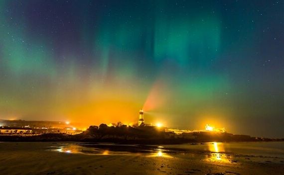 Aurora borealis vult de nachthemel met groene en blauwe tinten boven een vuurtoren op een rondreis Ierland. De sterrenhemel vult de gloed aan. Beneden is het silhouet van de kustlijn zichtbaar, met warme lichten van gebouwen die op het water reflecteren, wat een serene sfeer creëert. | Echt Ierland Vakanties