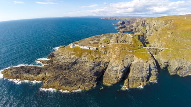 Luchtfoto van een ruige kustklif in Ierland met een historisch bouwwerk op de top, omgeven door een diepblauwe zee. Deze prachtige plek is perfect voor een avontuurlijke Ierland vakantie. De lucht is gedeeltelijk bewolkt en je kunt zelfs ver land aan de horizon zien tijdens je rondreis Ierland. | Echt Ierland Vakanties