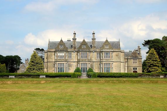 Een groot, sierlijk landhuis met meerdere schoorstenen en steile, puntdaken staat tegen een gedeeltelijk bewolkte lucht. Deze perfecte stop op een rondreis Ierland heeft hoge, smalle ramen, geflankeerd door verzorgde heggen. Op de voorgrond is een uitgestrekt gazon te zien dat naar het statige gebouw leidt. | Echt Ierland Vakanties