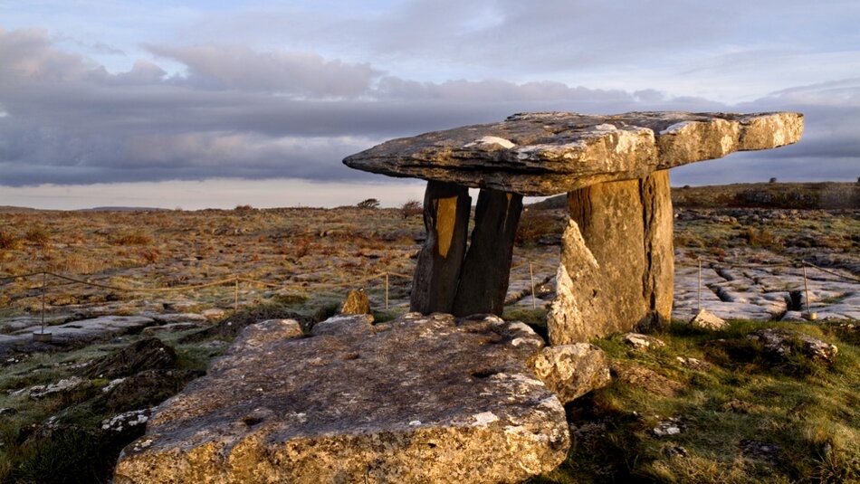 Een oude stenen dolmen staat in een grasachtig, rotsachtig landschap bij zonsondergang, wat een hint geeft naar de magie van een rondreis Ierland. Grote platte stenen worden ondersteund door rechtopstaande stenen, waardoor een tafelachtige structuur ontstaat. De bewolkte lucht laat een straaltje zonlicht door, wat een warme gloed werpt op dit iconische tafereel van uw Ierland vakanties. | Echt Ierland Vakanties