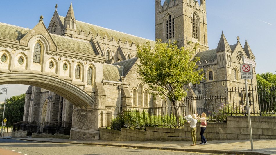 Twee mensen zwaaien onder een boom bij een historische stenen kerk met gotische architectuur, puntige bogen en een vierkante toren op hun rondreis Ierland. Een weg met een bordje 'niet parkeren' loopt langs het omheinde kerkterrein, terwijl de lucht helder en blauw blijft. | Echt Ierland Vakanties