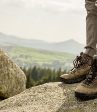 Close-up van een persoon die versleten bruine wandelschoenen en beige broek draagt op een grote rots, tegen de achtergrond van groene heuvels en verre bergen. De bewolkte lucht roept een gevoel van avontuur op, perfect voor degenen die dromen van rondreis Ierland of Ierland vakanties. | Echt Ierland Vakanties