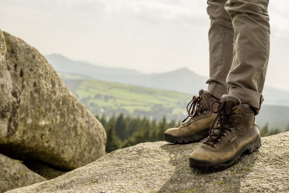 Close-up van een persoon die versleten bruine wandelschoenen en beige broek draagt op een grote rots, tegen de achtergrond van groene heuvels en verre bergen. De bewolkte lucht roept een gevoel van avontuur op, perfect voor degenen die dromen van rondreis Ierland of Ierland vakanties. | Echt Ierland Vakanties