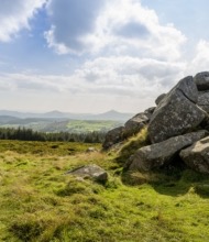 Een schilderachtig landschap in Ierland met grote, ruige rotsen aan de rechterkant, afstekend tegen een uitgestrekte vlakte van groene velden. Een rij bomen staat op de middengrond, met verre heuvels onder een gedeeltelijk bewolkte blauwe lucht. Perfect voor degenen die op zoek zijn naar rust tijdens hun vakantie in Ierland. | Echt Ierland Vakanties