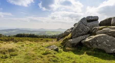 Een schilderachtig landschap in Ierland met grote, ruige rotsen aan de rechterkant, afstekend tegen een uitgestrekte vlakte van groene velden. Een rij bomen staat op de middengrond, met verre heuvels onder een gedeeltelijk bewolkte blauwe lucht. Perfect voor degenen die op zoek zijn naar rust tijdens hun vakantie in Ierland. | Echt Ierland Vakanties