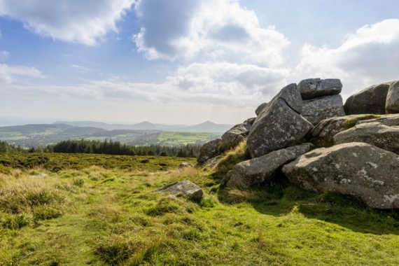 Een schilderachtig landschap in Ierland met grote, ruige rotsen aan de rechterkant, afstekend tegen een uitgestrekte vlakte van groene velden. Een rij bomen staat op de middengrond, met verre heuvels onder een gedeeltelijk bewolkte blauwe lucht. Perfect voor degenen die op zoek zijn naar rust tijdens hun vakantie in Ierland. | Echt Ierland Vakanties