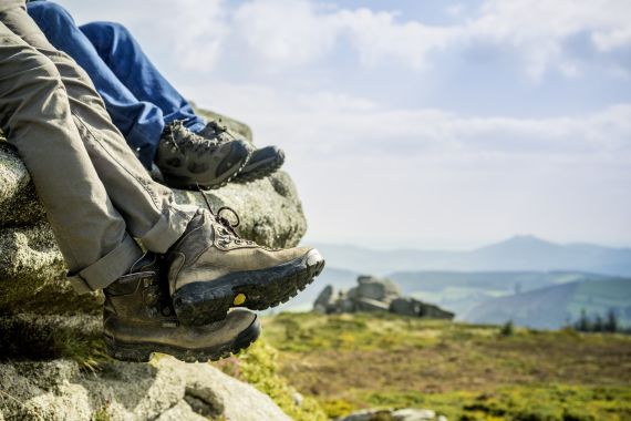 Twee mensen zitten op een rotsachtige richel, laarzen klaar voor avontuur. Het ene paar is donkerbruin met gele details, het andere zwart met grijze accenten. Achter hen, glooiende heuvels en een gedeeltelijk bewolkte lucht hinten naar een onvergetelijke vakantie Ierland ervaring in buitenpracht. | Echt Ierland Vakanties